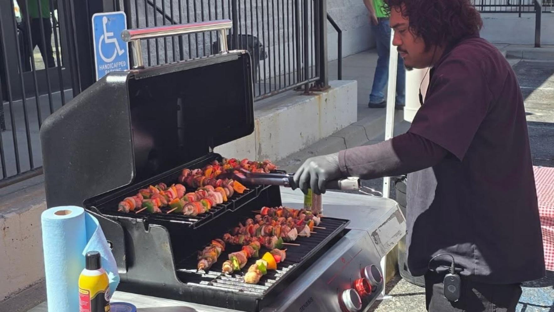 A man cooks colorful meat and vegetable skewers on an outdoor gas grill.
