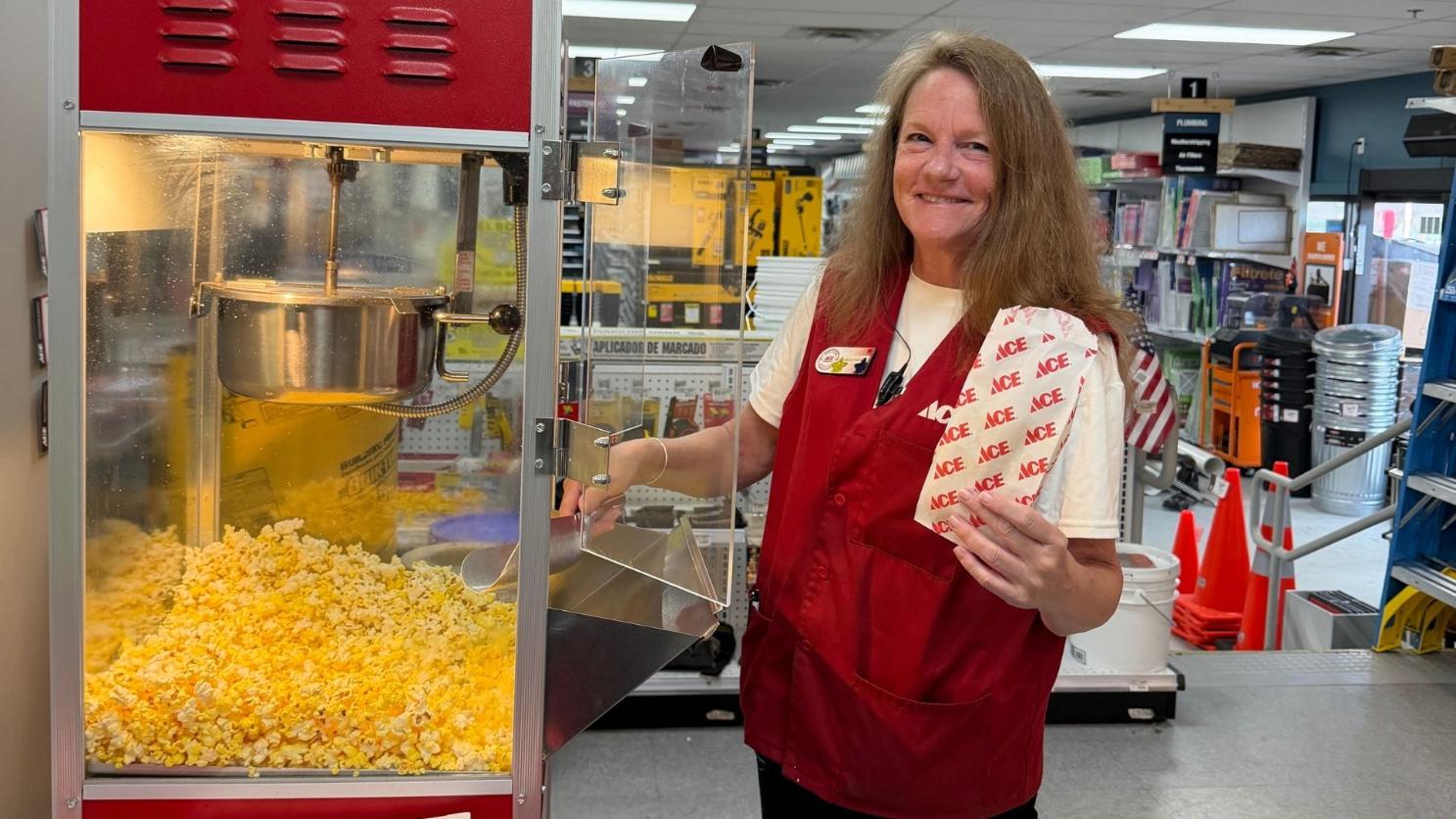 Smiling Ace Hardware employee holds a bag of fresh popcorn beside a popcorn machine.